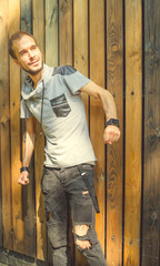 A young handsome man of slim build in casual clothes is posing against a wooden fence.