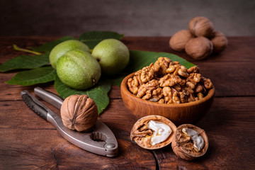 Walnuts chopped in a bowl, whole and in a green peel and a nut cracker on a dark old wooden background.