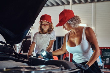 Mother is teaching her daughter how to fix broken car's engine at workshop.