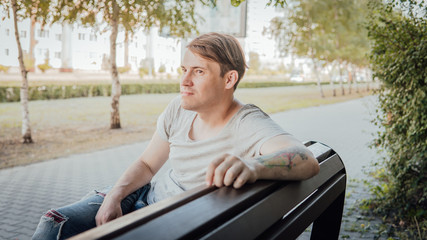 A young handsome man in casual clothes sits on a bench in a small alley.