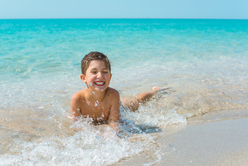 A cheerful kid on the beach lies in the clear sea water.