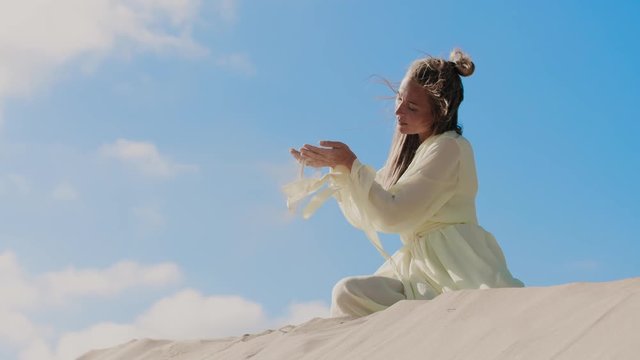 Woman in light yellow dress on sand dune in fornt of blue sky pouring sand from hands on wind. Slow motion