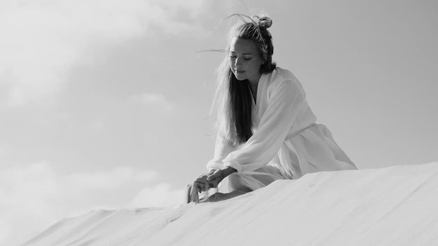 Woman in light dress on sand dune in fornt of sky pouring sand from hands on wind. Slow motion black and white