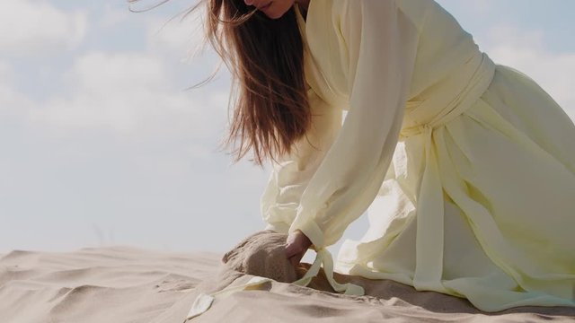 Woman in light yellow dress on sand dune in fornt of blue sky pouring sand from hands on wind. Slow motion