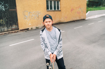 Fototapeta premium Teenager skateboarder boy in baseball cap street portrait holding skateboard on asphalt road near a grunge graffiti wall. Youth generation Freetime spending concept image.