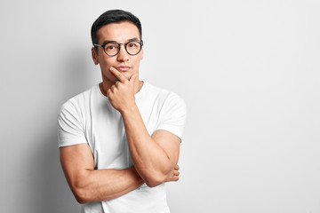 Smart Kazakh dressed in casuals and glasses thinks and doubts, holds his chin with hand on white studio background. Responsible Asian makes a decision
