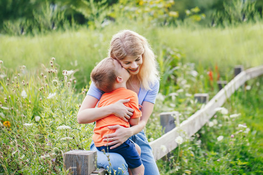 Young blonde Caucasian mother and boy toddler son hugging kissing in park outdoor on summer day. Mom and child love and tenderness. Happy family childhood lifestyle.