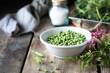 Young green peas in white bowl on wooden background. Pea flowers and daisy flowers on the table.