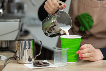 Barista in an apron pours milk into a green eco-friendly paper cup. Barista work in a coffee shop