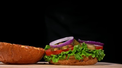 Craft burger is cooking on black background. Consist: sauce, lettuce, tomato, red onion, pickle, cheese, bacon, air bun and marbled meat beef.