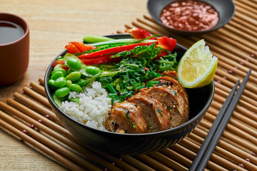 Delicious Buddha bowl with teriyaki chicken, fresh vegetables, edamame soy beans and basmati rice with green tea on a wooden table.
