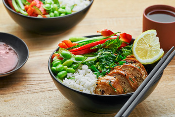 Wholesome meal made of teriyaki chicken fillet, fresh cucumber, red bell pepper, edamame, stew cabbage and basmati rice. Two delicious Buddha bowls with green tea cups on a table.