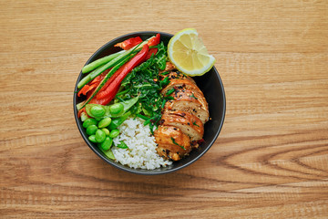 Teriyaki chicken fillet, fresh cucumber, red bell pepper, stew cabbage, edamame and basmati rice in black bowl. Traditional Asian Buddha bowl meal. Top view, directly above shot.
