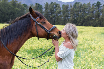 Woman kissing her chestnut Arab horse  on his nose, standing facing each other, outdoors with field of yellow flowers.