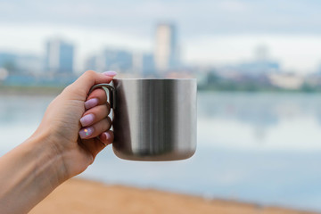 hand holding a metallic cup of coffee or tea. morning sunlight. drinking coffee near lake in city park. sunrise time.