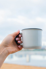 Obraz premium hand holding a white cup of coffee or tea. morning sunlight. drinking coffee near lake in city park. sunrise time.