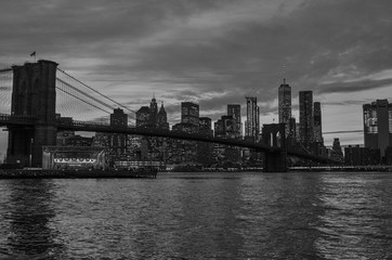 Fototapeta na wymiar Black and white photo of the Brooklyn Bridge during a cloudy sunset in foreground with New York City skyline in background.