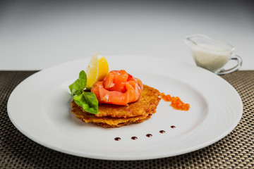 A horizontal photo of potato pancakes with salmon and red caviar, garnished with basil and lemon, served on a white plate.