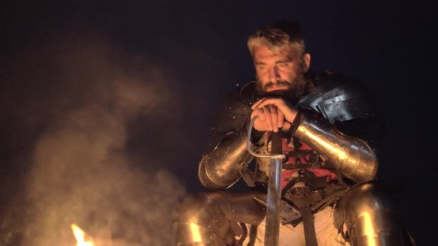 Portrait of medieval knight in armor whit sword sitting near the bonfire.