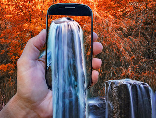 digital composite of a hand with a smart phone and a waterfall in autumn