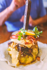 Whole butter roasted cauliflower, with garlic, melting brie and crispy bacon on a white plate with garnish and a knife with a wooden handle in the centre. A man can be seen ready to eat in soft focus.