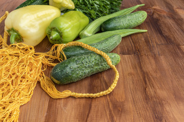 Green vegetables of zucchini, okra, cucumbers, bell pepper and a bunch of parsley lie next to a yellow string bag on a brown wooden background, side view