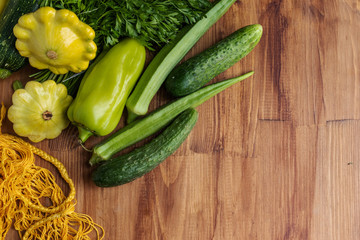 Green and yellow vegetables from zucchini, okra, cucumbers, bell pepper, a bunch of parsley and squash lie next to a yellow string bag on a brown wooden background, top view