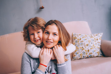 Closeup portrait of a little son and mom. Son hugs mom by the neck. Mom and son are looking into the frame and smiling.