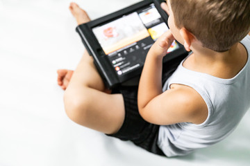 Little boy (child) sits on a white floor and watching cartoons on a tablet