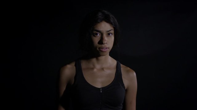 MS mixed race woman walks forward from soft focus against black background. She turns and comes into focus, looking at camera HD