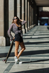 Beautiful young woman with longboard on the city street in sunny weather. Young hipster girl posing with skateboard.