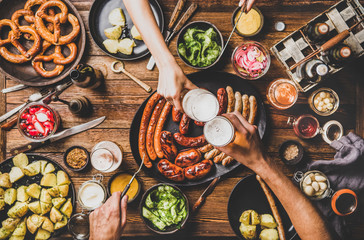 Flat-lay of Oktoberfest dinner table with grilled meat sausages, pretzel pastry, potatoes, cucumber...