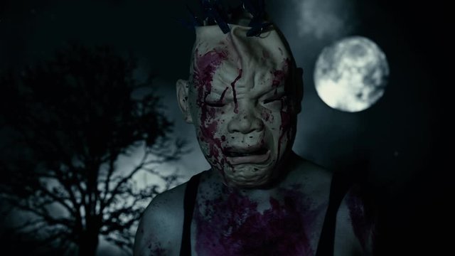 A scary man in a bloody mask in a cemetery, against the backdrop of a full moon and dead tree in dusk.. Horror character concept. Scary places. Halloween background