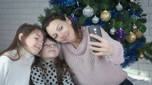 The family takes a selfie. Happy mother with her daughters is photographed on the phone against the background of a Christmas tree.