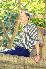 Young American Man relaxing outside in New York City, wearing black, white striped, long sleeve shirt, blue pants, wristwatch, sitting on top of wall by flowerpot in hot summer, frowned, sad, thinking