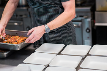 male chef preparing to go orders in industrial kitchen, wearing a watch and a striped apron, serving enchiladas out of a large baking dish
