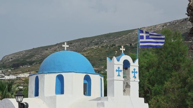 Traditional Greek Orthodox church in Parikia town. Paros island, Greece. Temple of christian religion in blue and white typical colors. Mediterranean vegetation. Wind blowing flag on ancient building