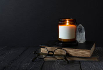 candle burning on stacked books, reading glasses, crystal, antique books, burning scented candle with blank label, copyspace, dark background with copy space, autumn mood, candle label design mock up