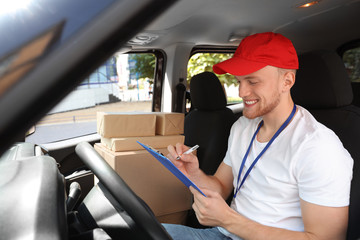 Young courier with clipboard and parcels in delivery car