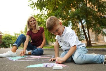 Nanny with cute little boy drawing house with chalks on asphalt