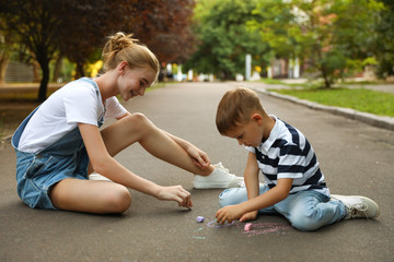 Teen nanny and cute little boy drawing with chalks on asphalt