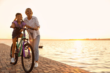 Fototapeta premium Happy mother teaching her daughter to ride bicycle near river at sunset