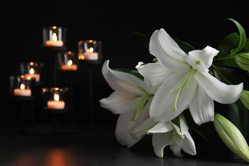 White lilies and blurred burning candles on table in darkness, closeup with space for text. Funeral symbol