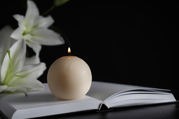 Burning candle, book and white lilies on table in darkness, closeup with space for text. Funeral...