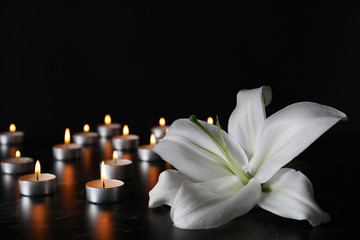 White lily and blurred burning candles on table in darkness, closeup with space for text. Funeral symbol