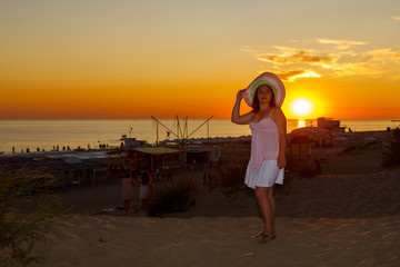 Woman in a white dress and a straw hat on a background of the sea at sunset.