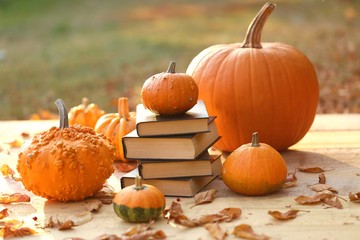 Autumn books. Halloween books. Stack of books with black cover and orange pumpkins set on a wooden table on a blurry background in bright sunshine.Cozy autumn mood. 