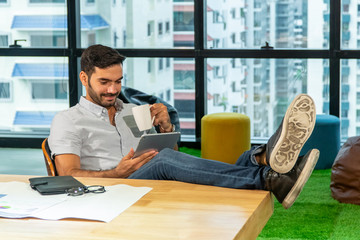 Relax young businessman sitting on the chair taking a break drinking coffee and using digital tablet for social media in office building. Smart office worker business man resting at modern workplace.