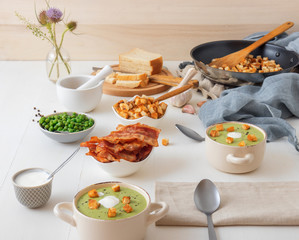 White wooden table with bowls of thick green pea soup embellished with roasted rusks, green peas, slices of grilled bacon, crushed black pepper and cream.