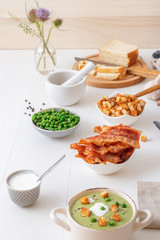 White wooden table with bowls of thick green pea soup embellished with roasted rusks, green peas, slices of grilled bacon, crushed black pepper and cream.